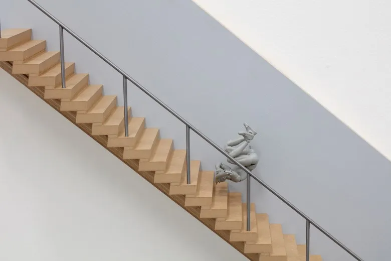 Close up of wall-mounted installation shows a miniature set of wooden stairs with a metal railing, and a grey kinetic figuring used for drawing mid-tumble down the stairs.