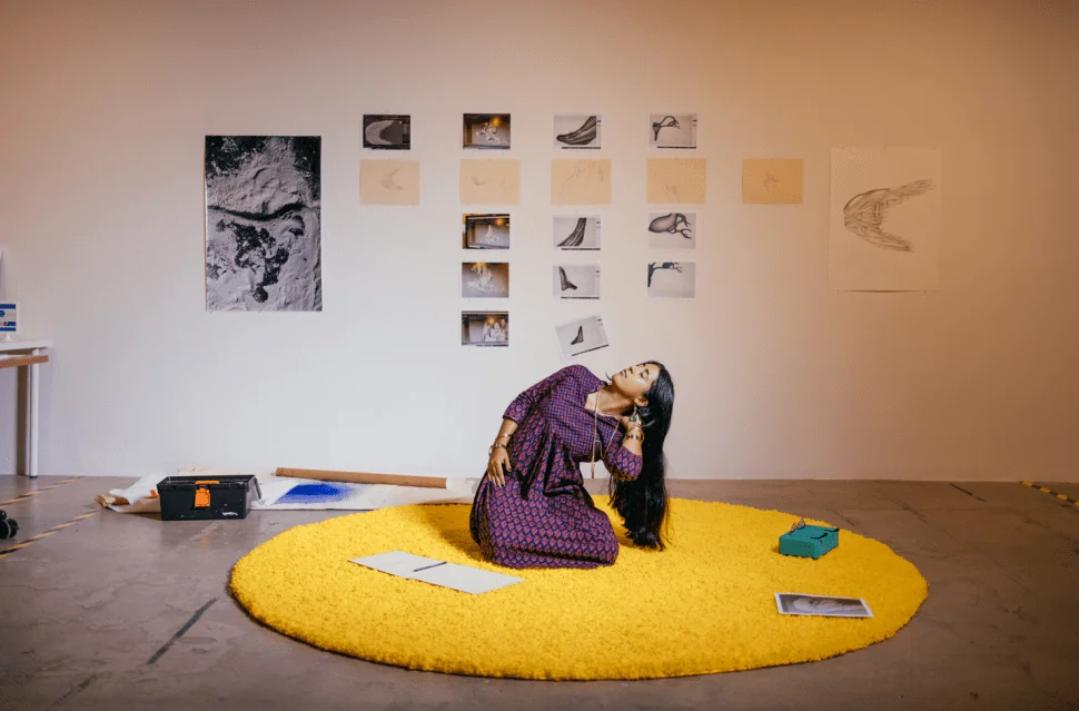 A photo of a woman sitting on a bright yellow circular rug in an art studio, surrounded by papers, with artwork hanging on the wall behind her. She is leaning sideways, looking up, while various sketches and photos are displayed on the wall.