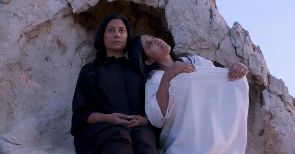 A photo of two women sitting together in front of a rock formation, one wearing black and looking straight ahead, and the other in white, leaning on the first woman with her head resting on her shoulder. They are seated in a relaxed manner, seemingly contemplative.