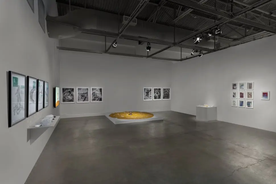 A photo of an art gallery with dim lighting, showing framed artwork on the walls, a circular installation in the center with yellow elements, and other small art displays. The gallery has a minimalist layout with an industrial ceiling and spotlights.