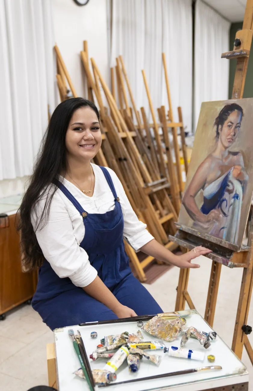 A woman sits on a easel with a portrait painting on it and paints surrounding her.