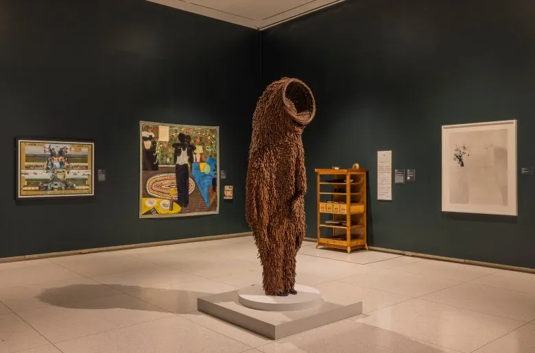 In the foreground, on a low plinth, stands a brown version of one of Nick Cave's Soundsuits, resembling a bear. In the back, on a dark gallery wall, are Kerry James Marshall's Slow Dance, featuring a couple dancing in a domestic setting, beside an abstracted wall work. To the right are a wooden shelf and a large white wall work.