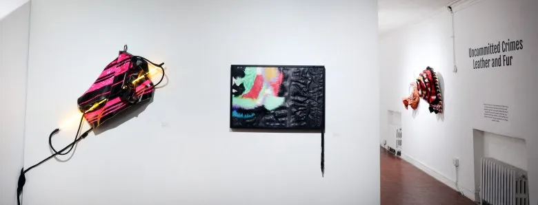 On the right wall, the exhibition title is displayed alongside colorful wall sculptures that are slightly out of focus. in the foreground are two colorful wall works. One has neon light through it while the other is rectangular with black and bright abstract shapes.