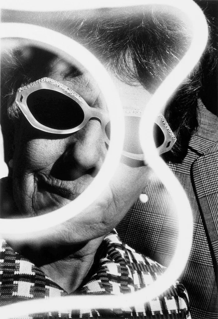 An older white women's face, with plastic, 60s-era sunglasses on, is seen behind another exposure of a wavy-shaped neon light. the image is in black and white, and the woman looks at the camera. She has short dark hair and wears a printed jacket.