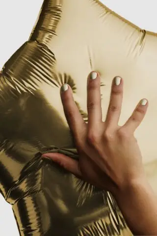 A hand holding a gold balloon wearing JW Anderson x Pleasing gold nail polish