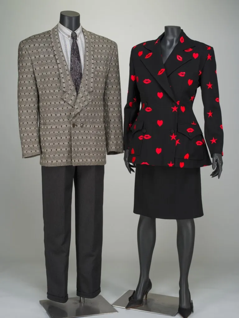 on the left is a mannequin wearing a men's suit, black pants and a beige jacket with a purplish tie and white shirt. on the right is a mannequin wearing a women's suit consisting of a black skirt and a black jacket with red lips and stars and hearts printed on it.