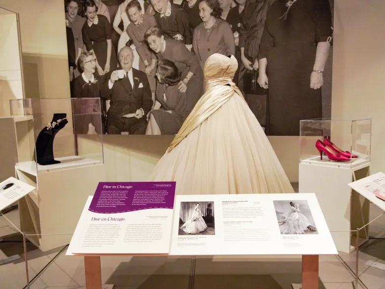 Installation view at Dressed in History exhibition shows a white strapless Dior gown displayed behind a placard about the designer. Beside the dress are a display of red high heels and a necklace. A black and white historical photo is on the wall behind the dress.