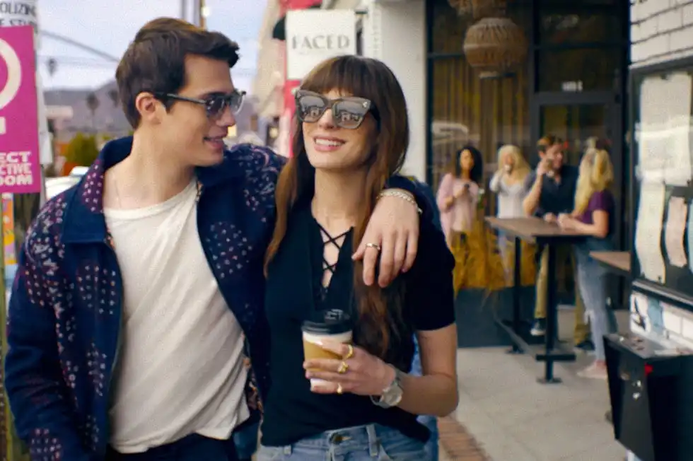 a couple walks arm in arm along a street with shops and people in the background