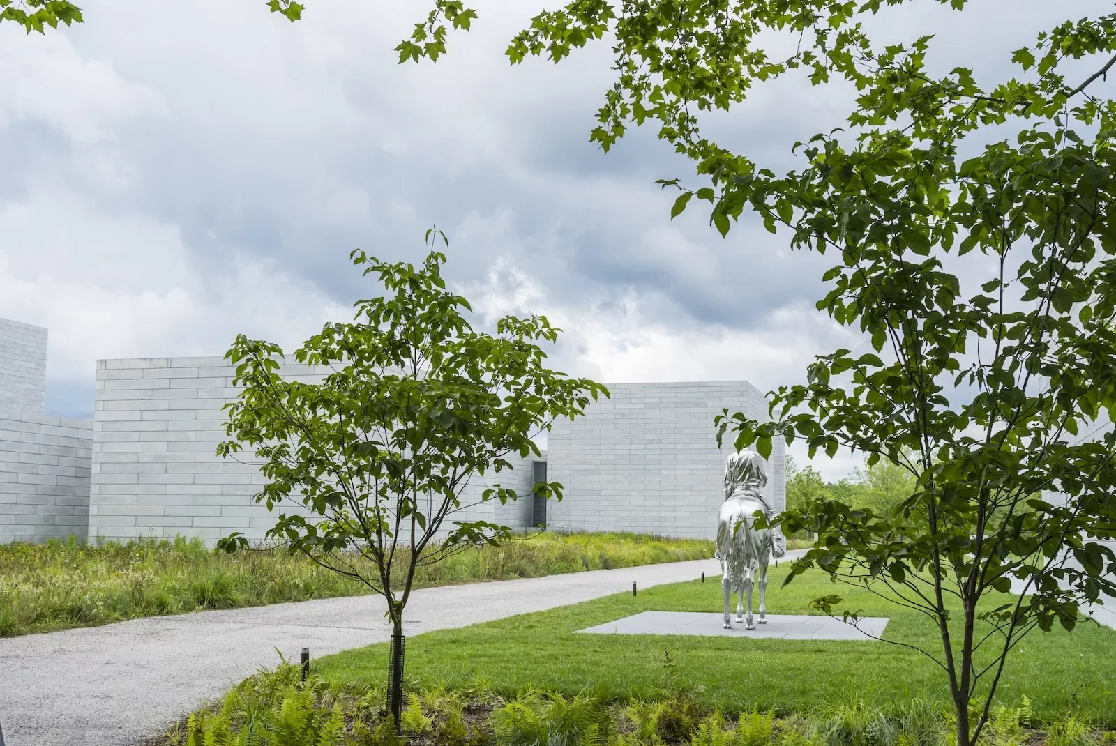 A lush green park space is seen on a deeply cloudy day, with a road and grass and trees and in the background, buildings made of white cut stones, and partly obscured by tree leaves, a shiny stainless steel horse and rider sculpture by Charles Ray.