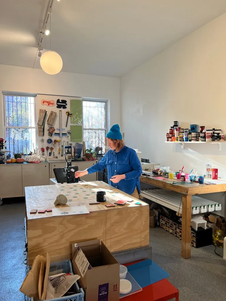 A color photo of a person standing in the makers area of Lot'sa, beside a large wooden table. Behind them on the walls are various tools and art supplies. Other supples can be seen on another wooden table behind them.