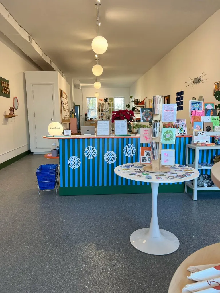 A view of Lot'sa shows the till, with a blue striped front. and various tables and shelves in the foreground stocked with prints, cards. and other small items.