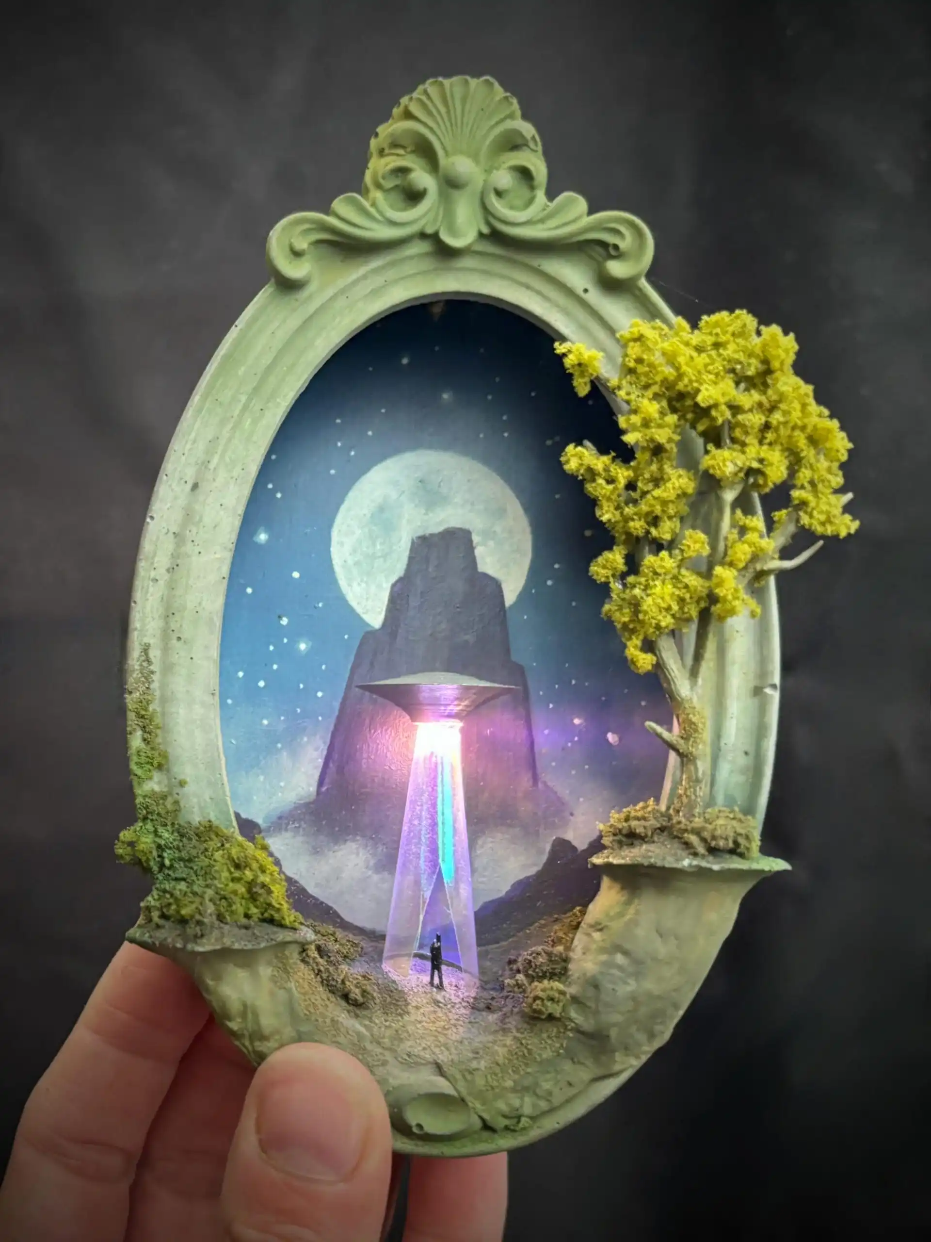 within a miniature ovoid frame, a detailed diorama depicts a landscape with a hovering UFO