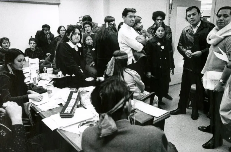 A black and white photo of a room full of Indigenous people sitting around and standing in the office of the BIA in Chicago. Some sit at desks while others crowd around them