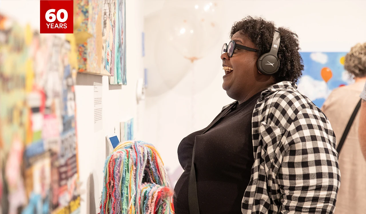 A woman stands in a bright and colourful art gallery looking at a painting, smiling and wearing headphones.