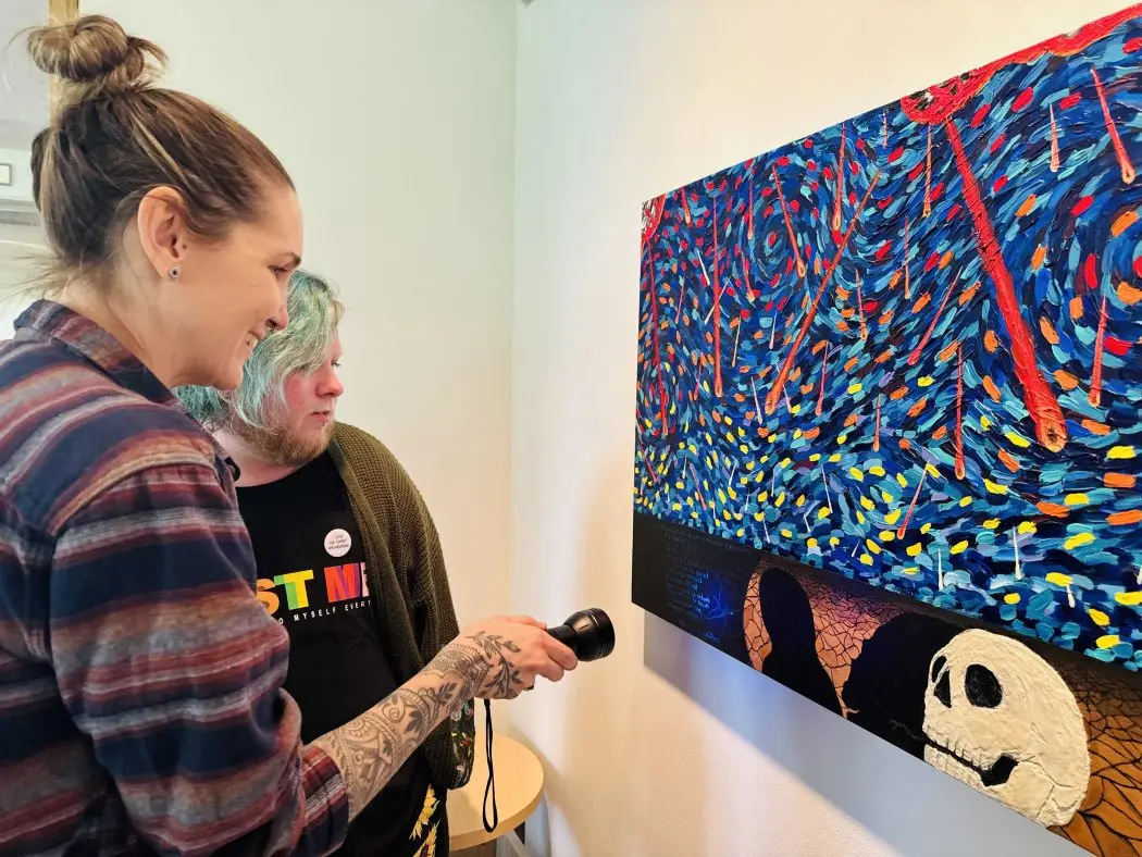 Two people stand together looking at a piece of art showing dark colours and images of skills, with one person shining a light on the painting to reveal hidden text.