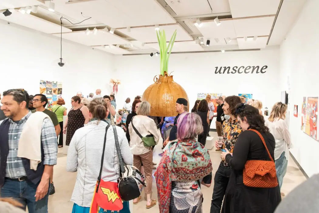 A group of people stand in a bright art gallery, socializing and looking at pieces of art.