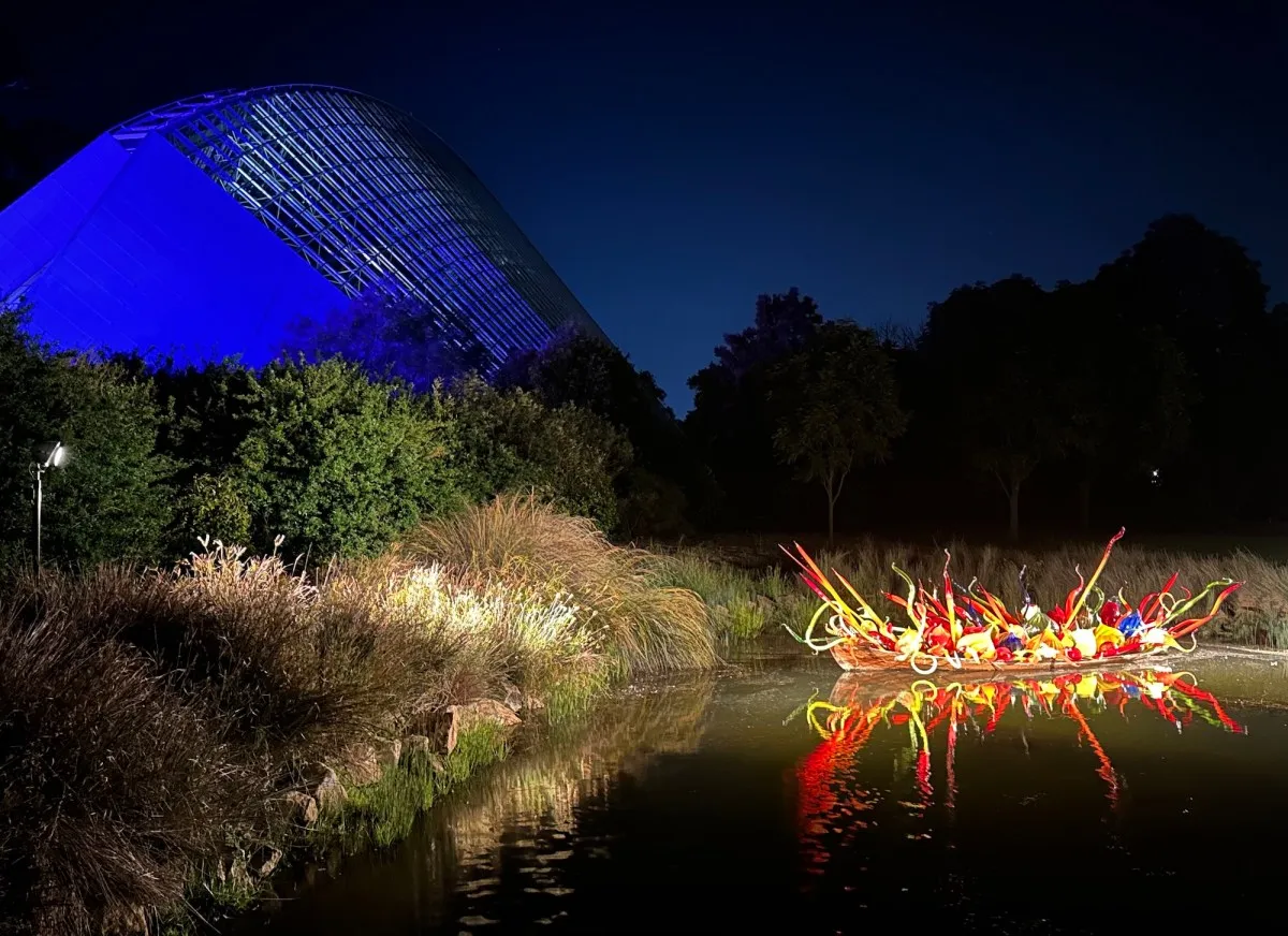 Night time view of an illuminated glass sculpture of a boat in garden setting. Dale Chihuly