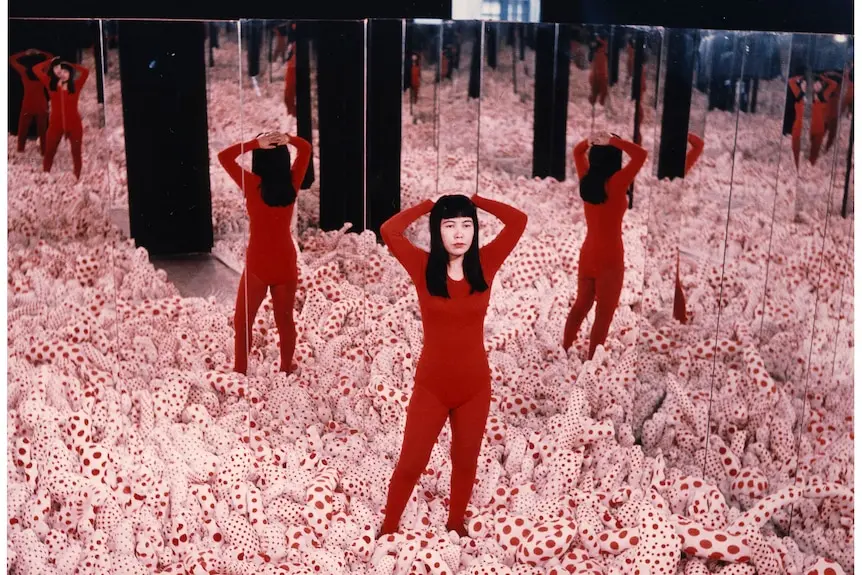 Yayoi Kusama, in red jumpsuit, stands in a room filled with smaller white and red objects, surrounded by mirrors.