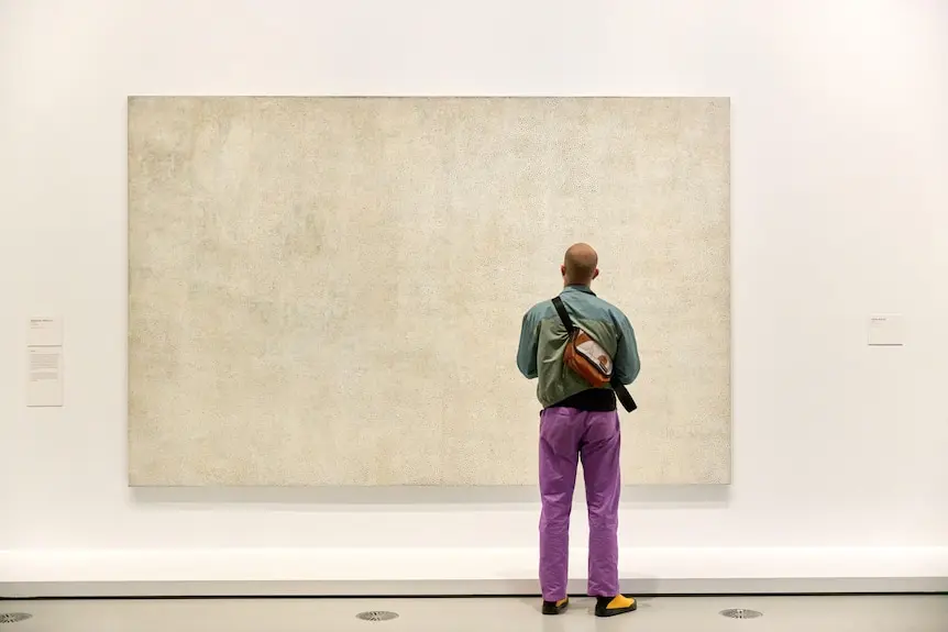 A person in purple pants stands in front of a large white canvas on a white wall.