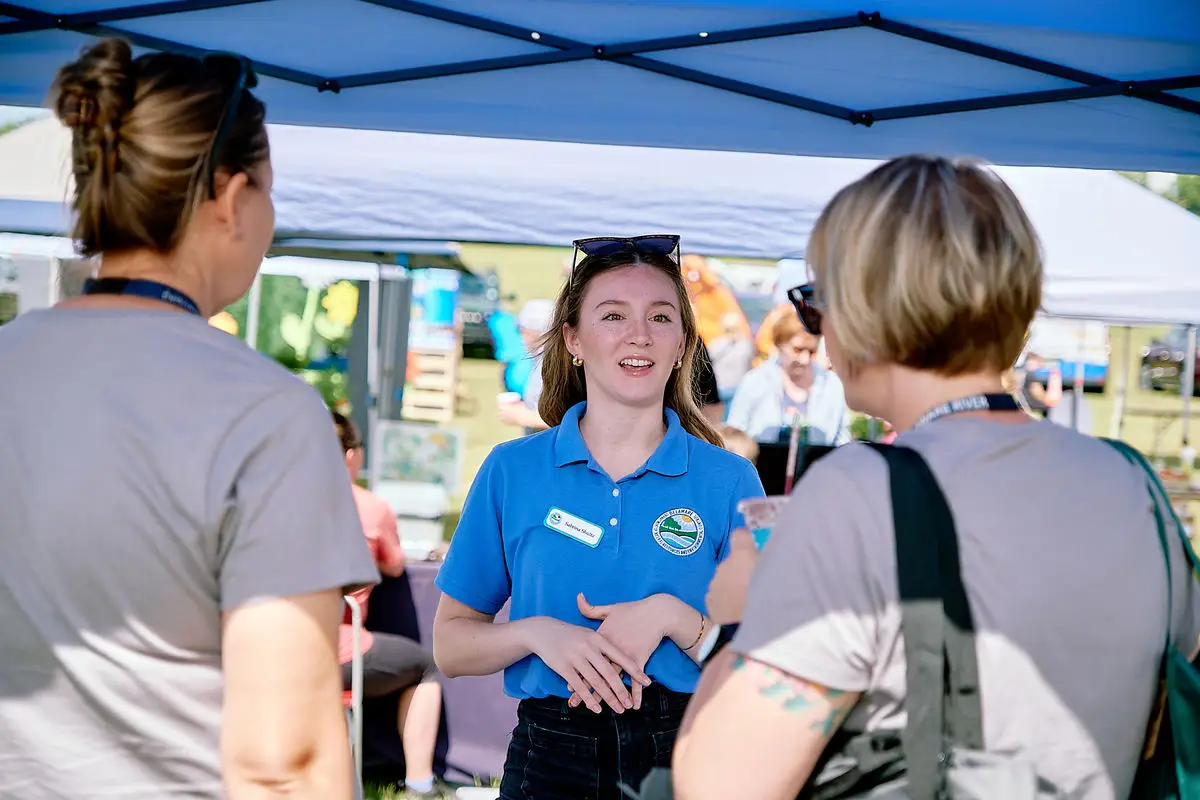 A state employee speaks with members of the public under a tent at an outdoor event.