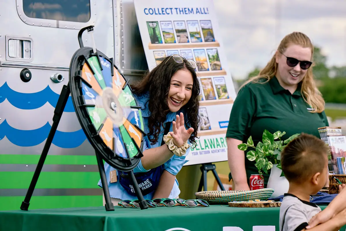 Two adults at a display table at an outdoor event smile as they talk to young children.