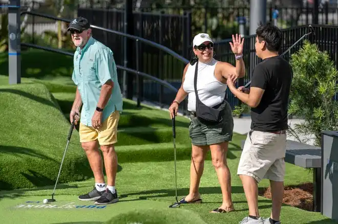 Daisy Leiva, center, gives John Mall a high five while playing golf with Perry Sigouin, left at PopStroke on December 20, 2024, in West Palm Beach, Florida. Tiger Woods, Greg Bartoliand TaylorMade Golf Company are the owners.