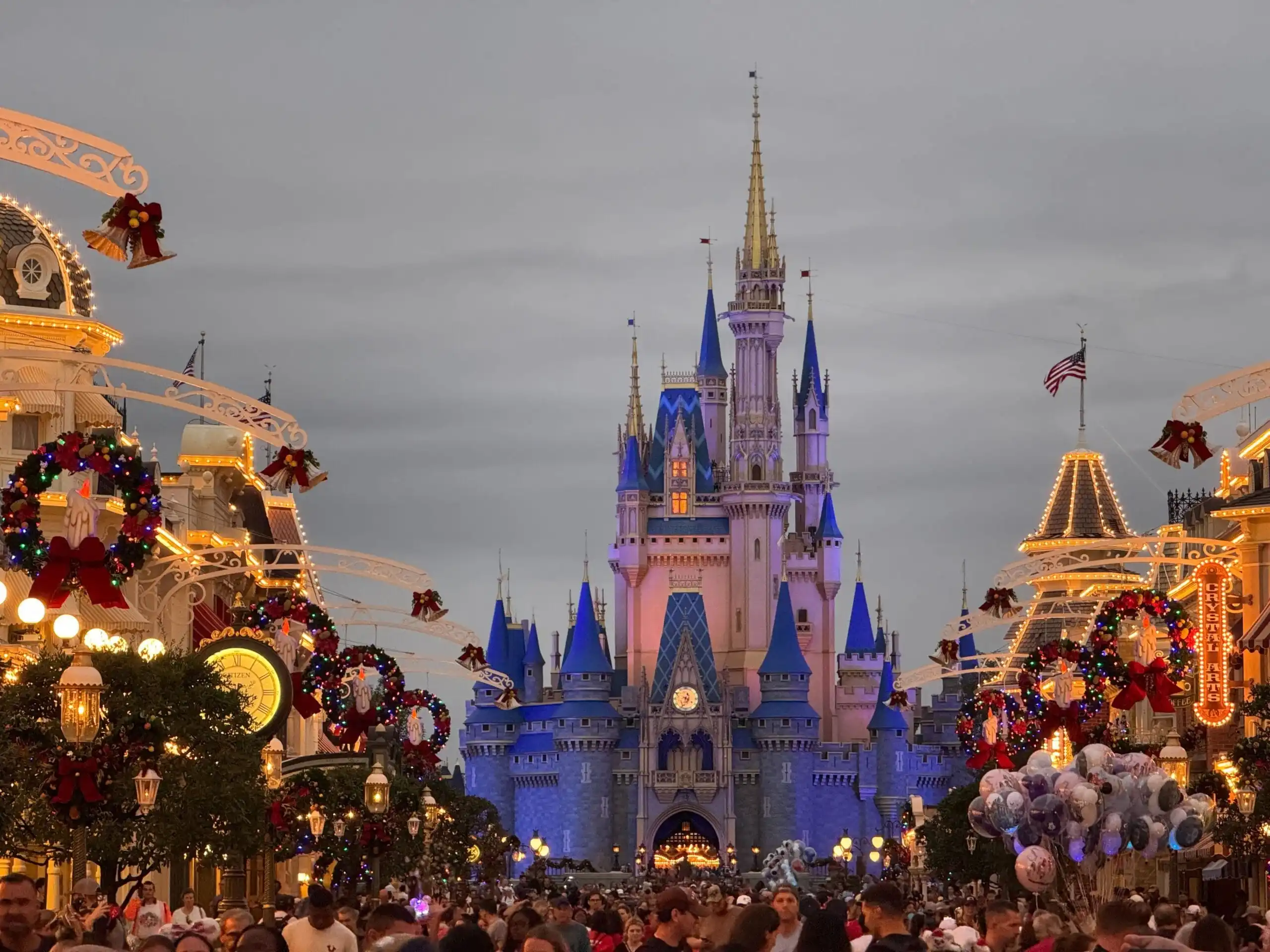 A large crowd gathers at a theme park main street, decorated with wreaths and lights. A fairytale castle is illuminated in the background. Lightning Lane Premier Pass