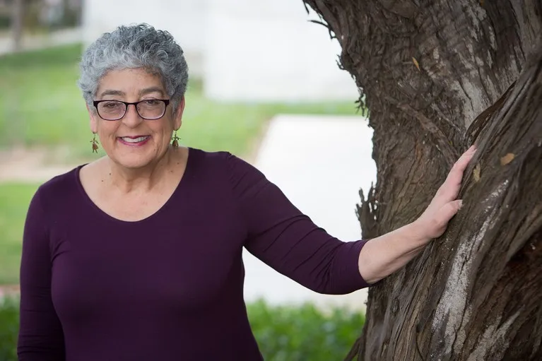 Portrait of Joanne Chory outside, holding onto a tree's trunk.