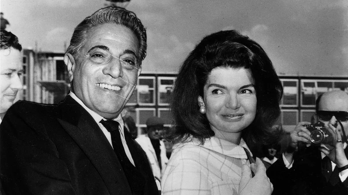 A black and white photo of Aristotle Onassis in a suit smiling next to Jackie Kennedy who looks at him from a side angle