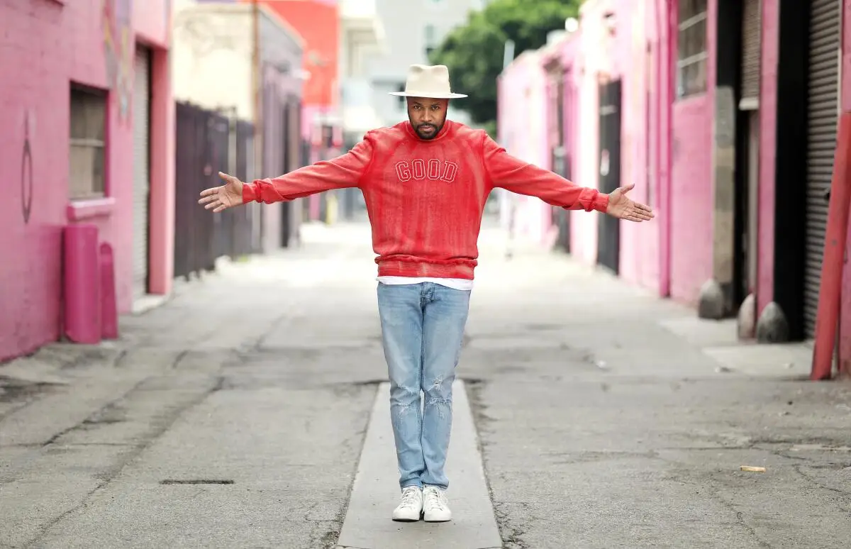 A man in white hat, red jacket and jeans stands, arms outstretched, with pink buildings on either side of him