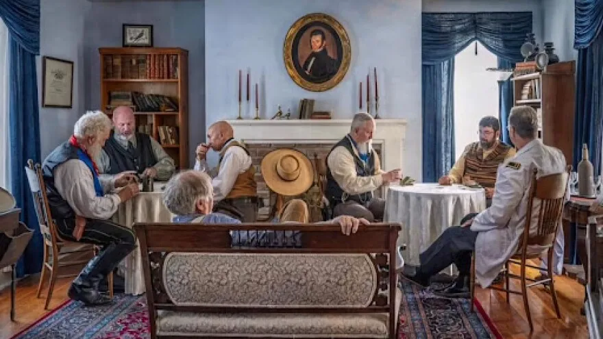 Seven men in period clothing sit in the parlor of a reconstructed 1800s home. 