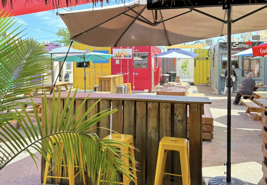 The outdoor seating area of the Iron Disrict in summer, with wooden counters, sun umbrellas, and yellow stools. 