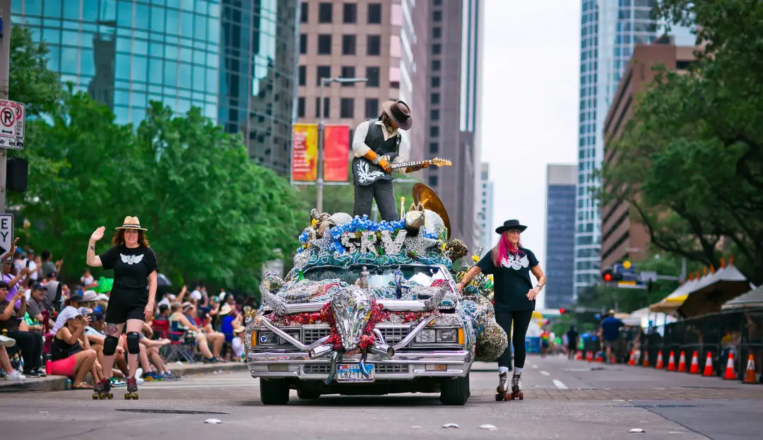 A parade featuring an art car and two people on either side of it.