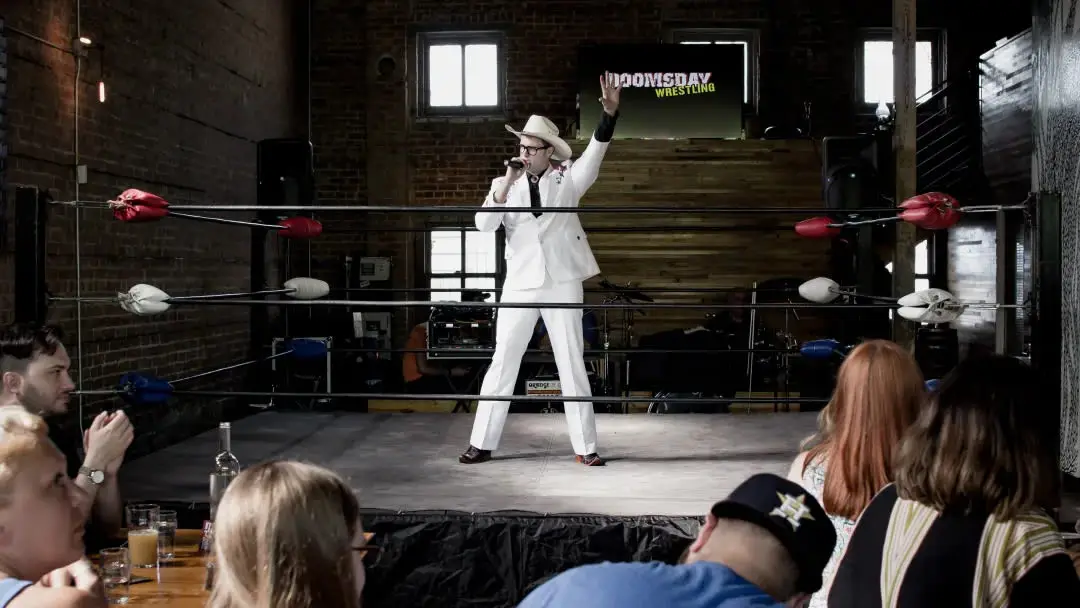 A man in a white suit and cowboy hat entertains a crowd from inside a wrestling ring.