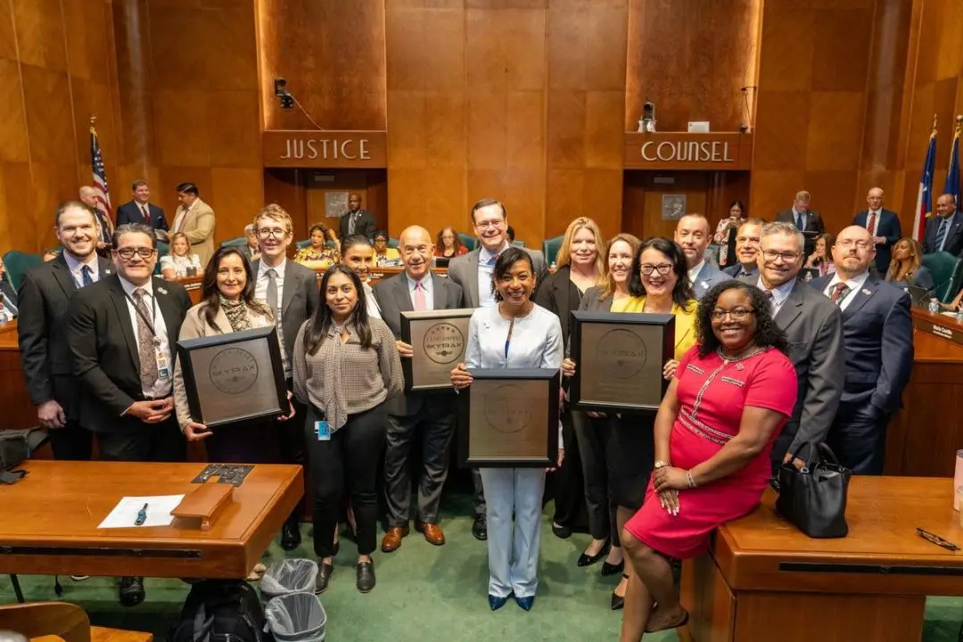 A large group of people in a serious-looking government office pose for the camera with four awards.