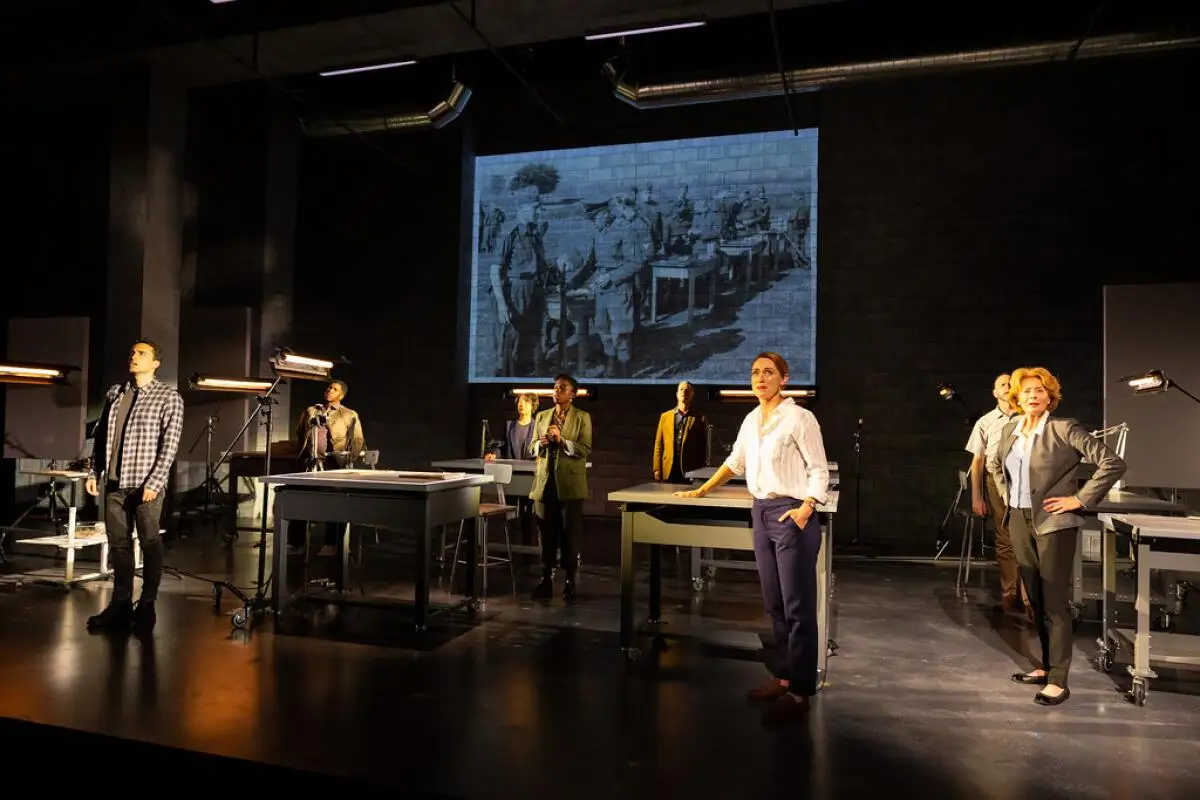 Onstage, people stand in a room with workbenches as a black-and-white film plays on a screen behind them