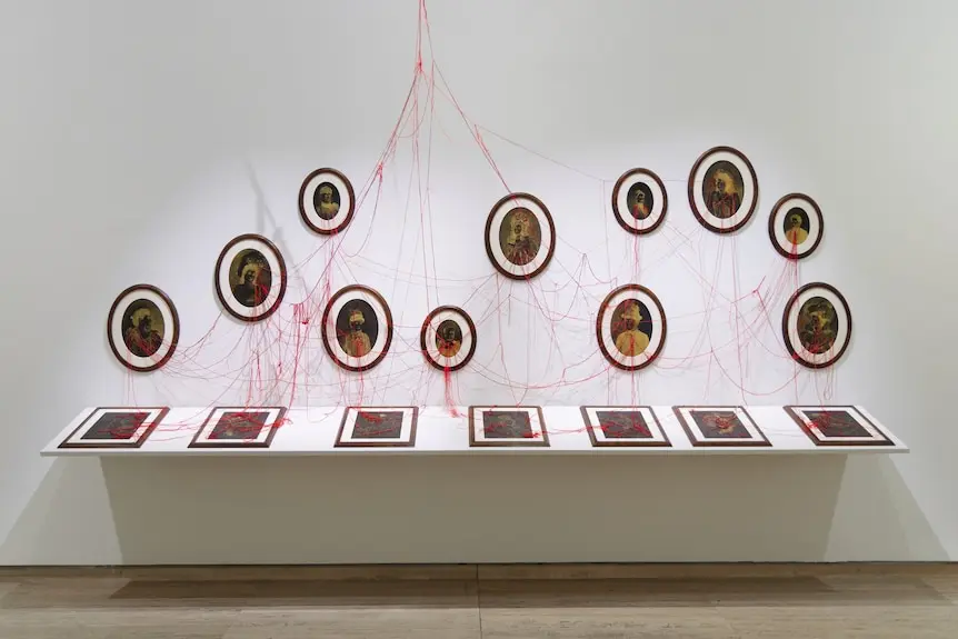 In a white room, a display cabinet shows several framed paintings just visible, and above it connected by red string are more.