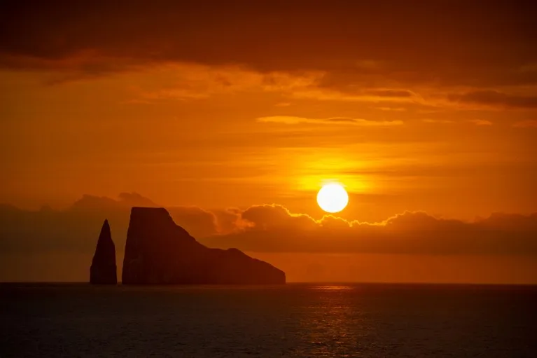 The sun sets behind two rock formations, lighting up the sky in orange