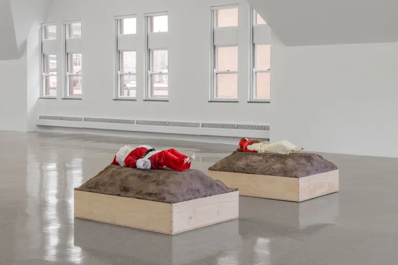 Two smaller than life-size humanoid sculptures, one dressed as Santa and the other as Jesus lay on dirt like platforms in the gallery, across from the Lincoln cabin. A set of six windows can be seen in the background.