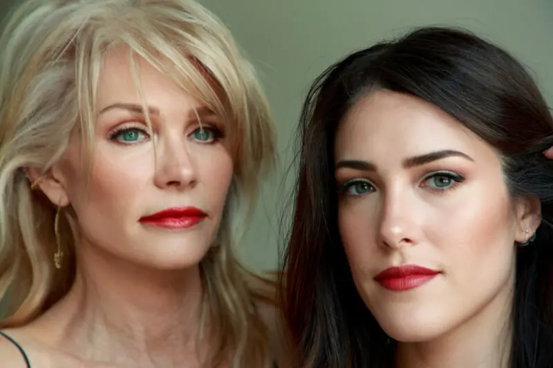 Two women with red lipstick are gazing at the camera. One has blonde hair and the other has dark brown hair.