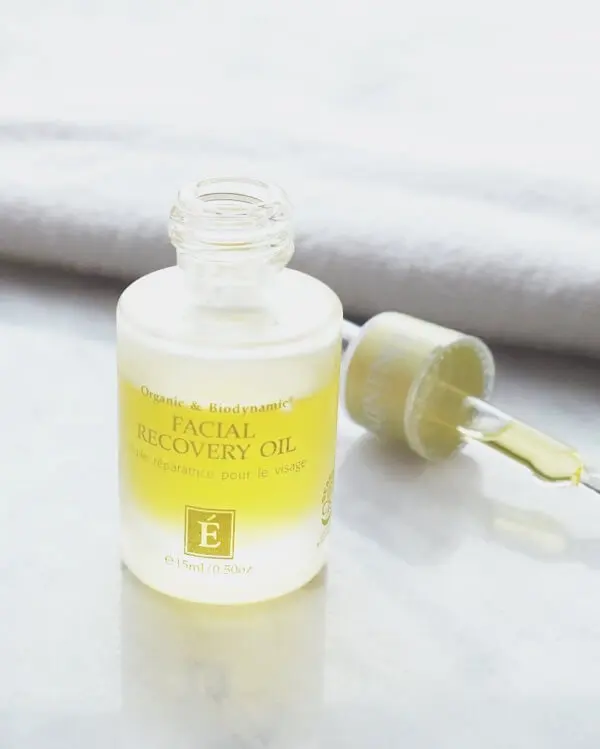 A bottle of Éminence Organic Skin Care Facial Recovery Oil with an open dropper sits elegantly on a light surface near a towel, showcasing the latest in expert skincare developments.