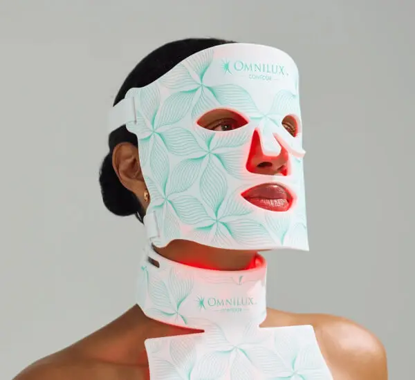 A person dons a full-face LED mask and neckpiece adorned with floral patterns, showcasing skincare developments. The mask emits a soothing red glow.