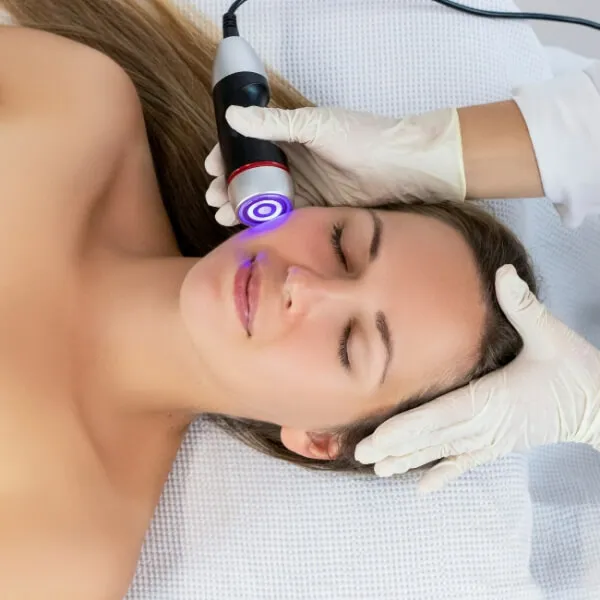 A person indulges in expert skincare, receiving a facial treatment with a handheld device emitting a blue light, meticulously applied by a gloved practitioner.