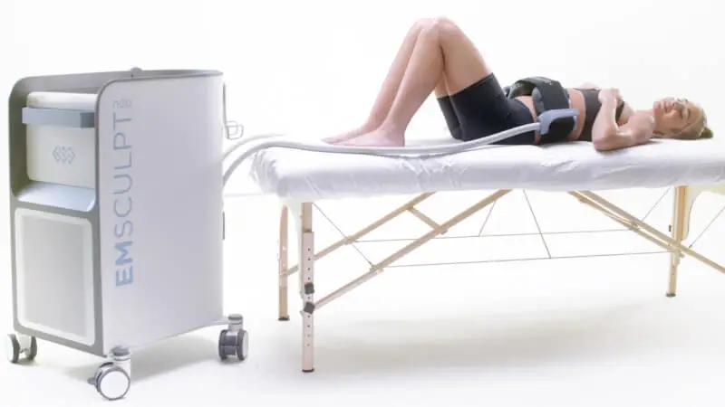 An expert technician oversees a session where a person lies on a therapy table with the advanced EMSCULPT Neo machine connected to their abdomen, highlighting the latest developments in skincare technology.