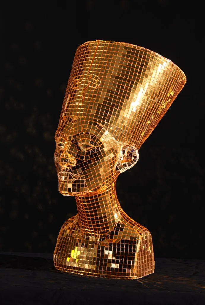 Gold mirror tiles cover a foam bust of Nefertiti. The bust faces to the left, with light reflecting off the side. The background is black.