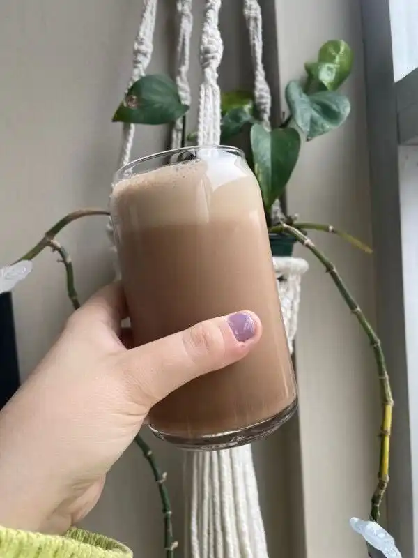 a hand holding a glass of brown beverage next to a plant