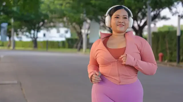 A young woman wearing peach-coloured jogging kit and cream headphones, jogs along a wide concrete path, lined by trees