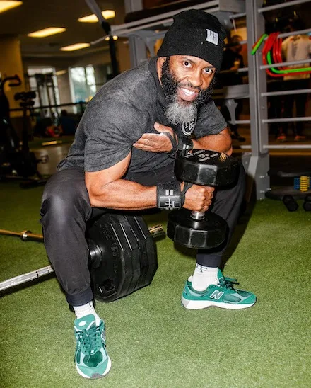 Jamar Blackshear, a Black man with a black and white beard, lifts a dumbell while seated on a barbell in a gym. He is wearing a Halal Fitness black beanie, a black t-shirt and wristbands and green New Balance sneakers.