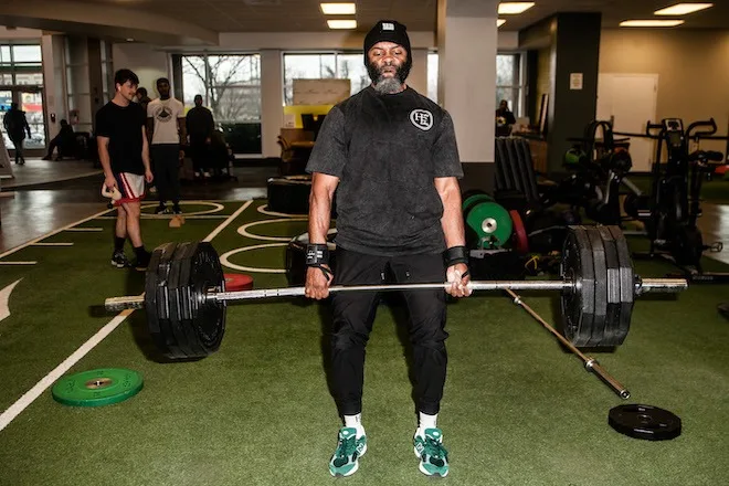 Jamar Blackshear, a Black man with a black and white beard, lifts a barbell in a gym. He is wearing a Halal Fitness black beanie, a black t-shirt and wristbands.