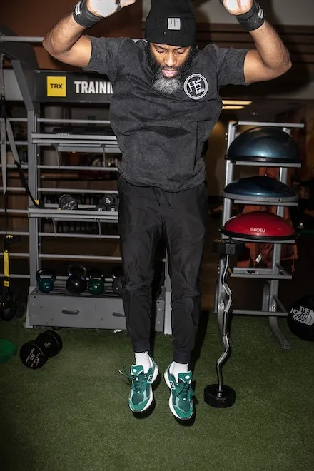 Jamar Blackshear, a Black man with a black and white beard, jumps while doing a burpee in a gym. He is wearing a Halal Fitness black beanie, a black t-shirt and wristbands and green New Balance sneakers.